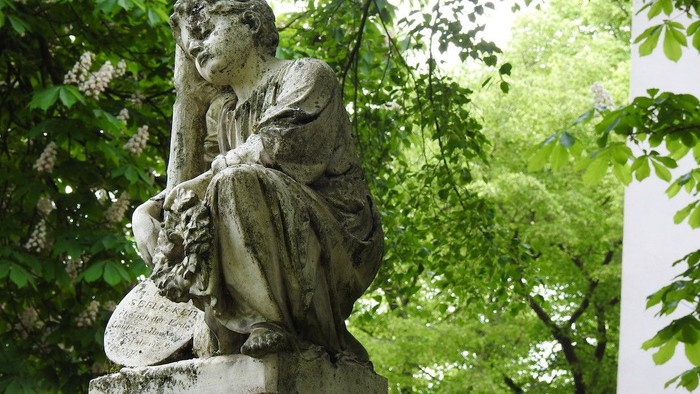 Eine friedvolle Steinstatue einer sitzenden Figur, die ein Buch hält, umgeben von üppigem grünem Laub.