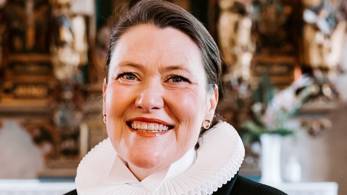 En kvinde i sort jakke med hvid krage står foran en prægtig altertavle.