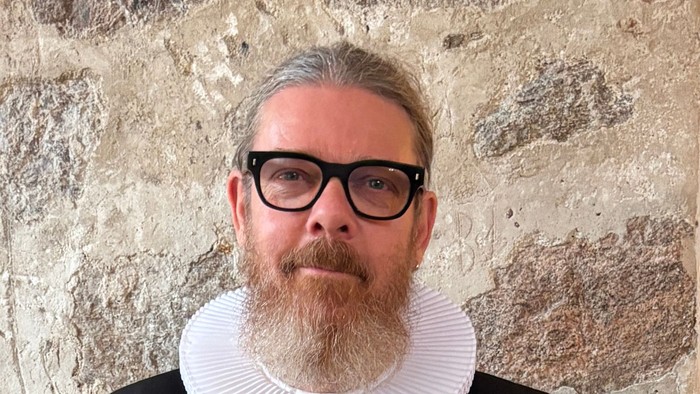 En mand i historisk tøj med en hvid kravekrøs, sort jakkesæt og briller poser foran en stenmur.