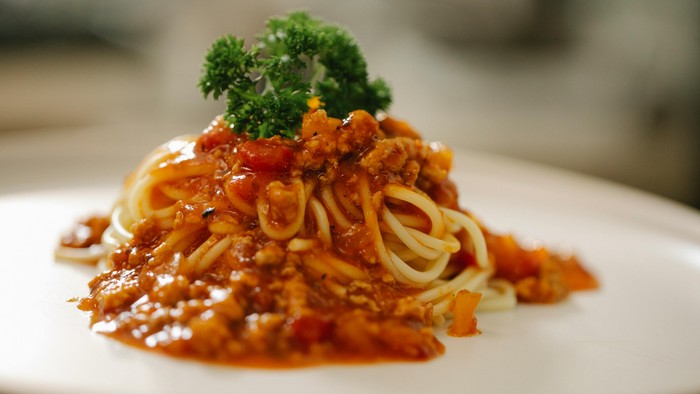 Spaghetti dækket af en fyldig kødsovs og toppet med en kvist persille på en hvid tallerken.