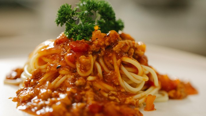 Spaghetti dækket af en fyldig kødsovs og toppet med en kvist persille på en hvid tallerken.