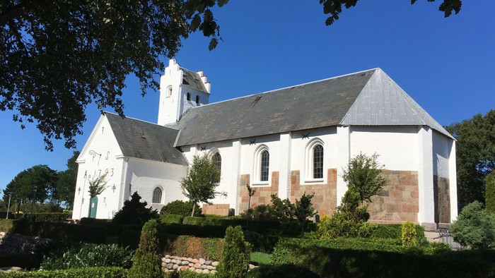 En lille, hvidkirurgisk kirke med tårn og bjergtegn i træ og mursten, omgivet af grønne planter og træer under et klarblåt himmel.