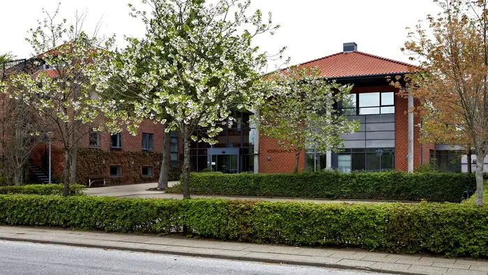 Moderne murstensbygning med glasvinduer, omgivet af veltrimmede hække og unge træer.