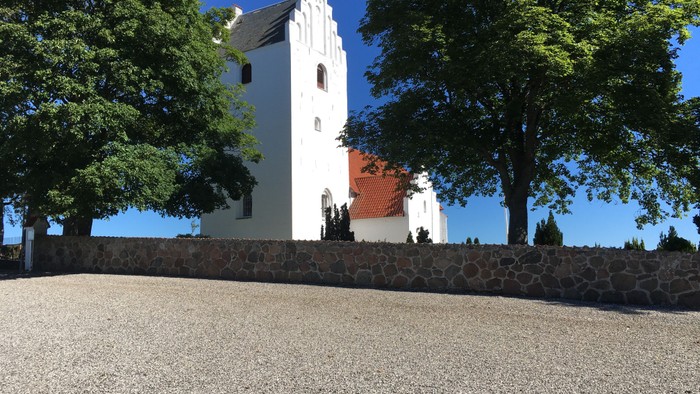 Et hvidt kirkebygning med tårn og røde dørme, omgivet af træer og en murstenge mur