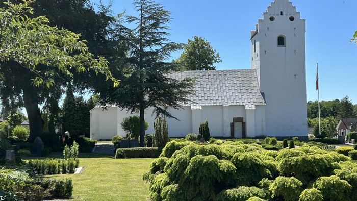 En fredfyldt hvid kirke med et højt spir står midt i frodig, velplejet grønne omgivelser under en klar blå himmel.