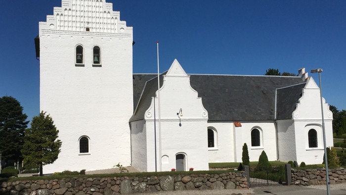 Hvid kirke med tårn og spids tak, omgivet af en mur og græsplæne