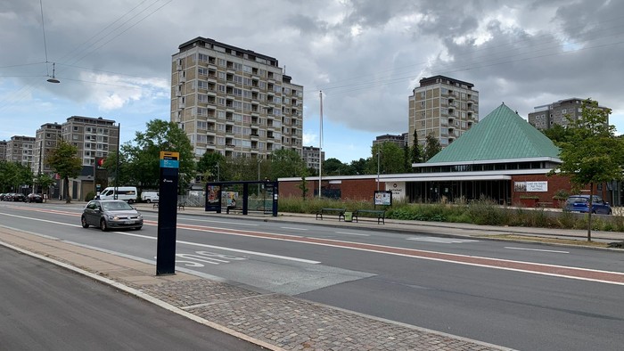 Bymæssig gadescene med højhuse, en busstoppested og en nærliggende struktur med grønt tag.