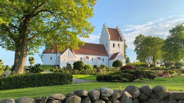 En smuk hvid kirke med rødt tag og en stor træ i forgrunden