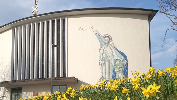 Moderne Kirche mit Wandgemälde eines Heiligen, umgeben von leuchtend gelben Osterglocken im Frühling.