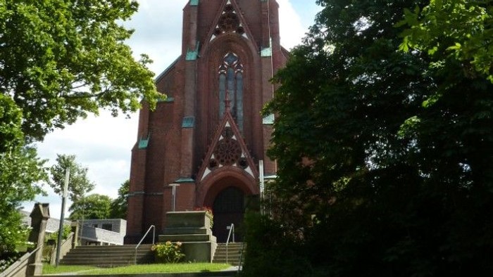 Kirchturm mit grünem Dach und Uhr, umgeben von Bäumen und Treppe davor