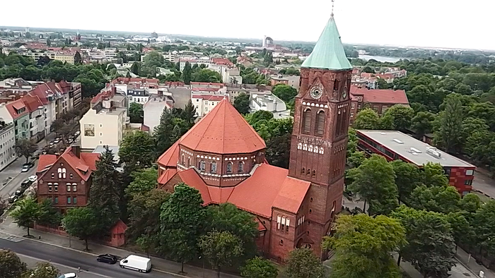 Luftaufnahme einer historischen Backsteinkirche mit hohem grünem Turm in einer grünen, städtischen Umgebung.