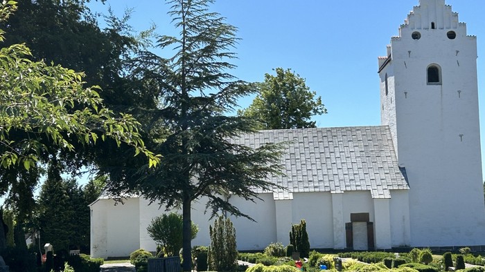 En fredelig hvid kirke med et højt spir omgivet af en frodig, grøn og velplejet have under en klar himmel.