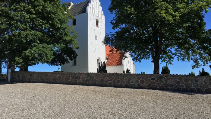 Et hvidt kirke med tårn og rødt tag bagved en murstenge mur og træer