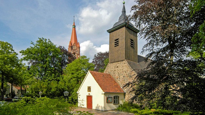 Kirche mit Uhrturm und kleinerem Gebäude davor, umgeben von Bäumen und Grünflächen.