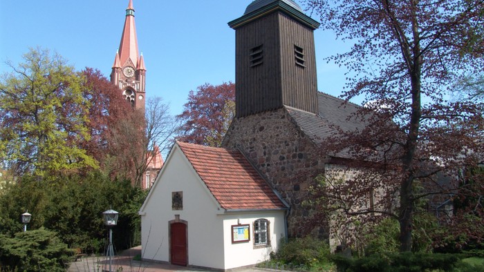 Idyllische Kirche mit einem kleinen weißen Gebäude und hohen Türmen, umgeben von herbstlichen Bäumen.