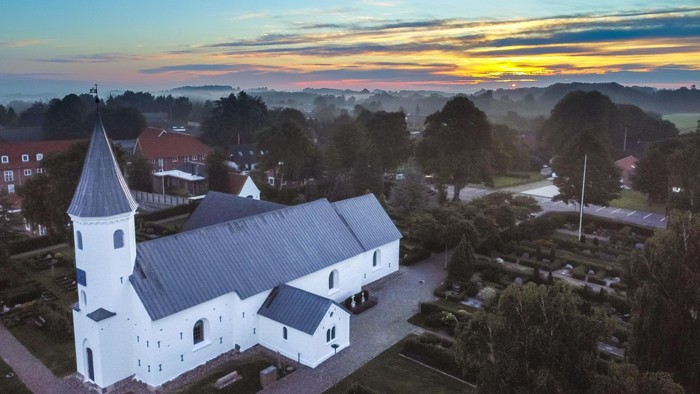 En fredelig kirke med hvid facade står midt i en rolig landsby ved solnedgang.
