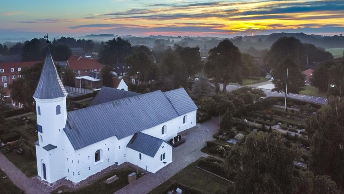 En fredelig kirke med hvid facade står midt i en rolig landsby ved solnedgang.