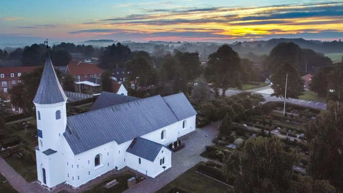 En fredelig kirke med hvid facade står midt i en rolig landsby ved solnedgang.