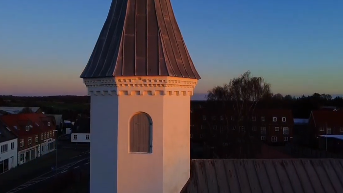 Et tårn badet i gyldent lys mod en tusmørkeby med tage og fjerne træer.