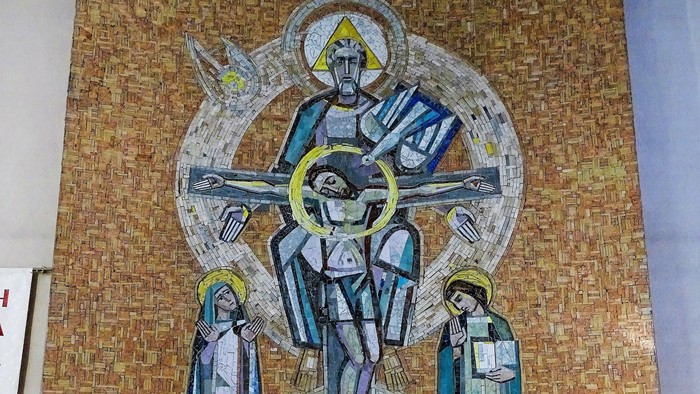 Ein Steinaltar mit Blumenschmuck und zwei Kerzen steht vor einem großen Mosaik-Wandbild. Das Mosaik zeigt zentral die Heilige Dreifaltigkeit.