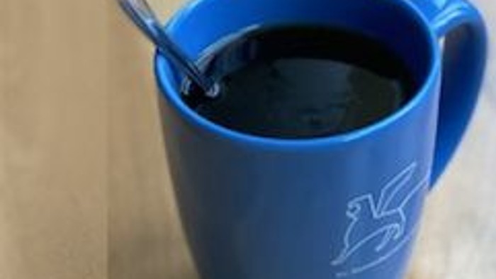 Eine blaue Tasse mit einem weißen Pferde-Logo enthält dunkle Flüssigkeit, begleitet von einem Löffel.