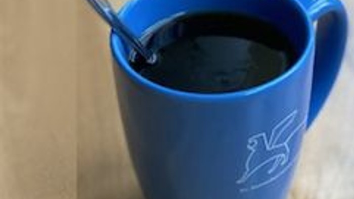 Eine blaue Tasse mit einem weißen Pferde-Logo enthält dunkle Flüssigkeit, begleitet von einem Löffel.