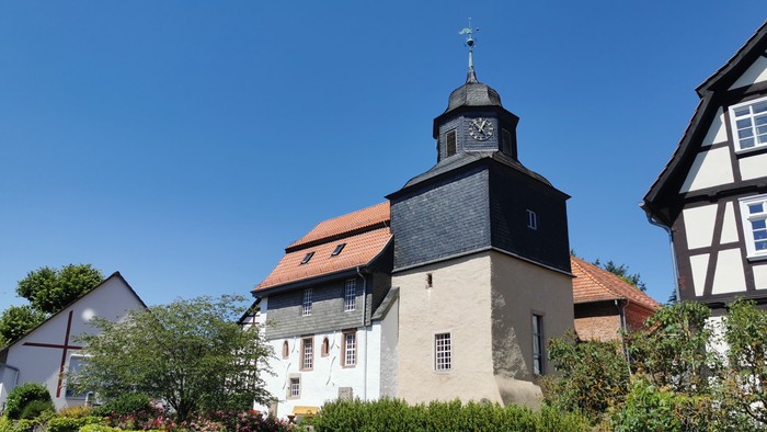 Das Bild zeigt eine Kirche mit schwarzem Dach und weißem Turm, umgeben von grünen Büschen und anderen Häusern.