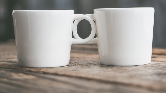 To hvide sammenkoblede krus med en cirkulær bro imellem, placeret på en træoverflade.