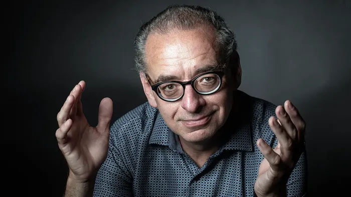 En mand med briller og hævede hænder, iført en mønstret skjorte på en mørk baggrund.