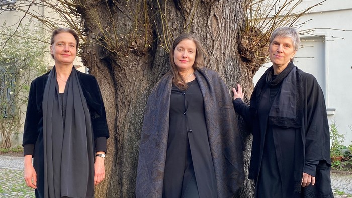 Drei Personen in schwarzer Kleidung posieren gemeinsam neben einem großen Baum im Freien.