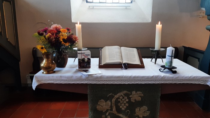 Eine ruhige Altargestaltung mit einem offenen Buch, Blumen, Kerzen und einer dekorativen Tischdecke auf einem antiken Tisch.