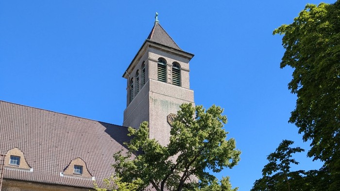Kirchturm mit Uhr und Kreuz, umgeben von Bäumen und Gebäuden