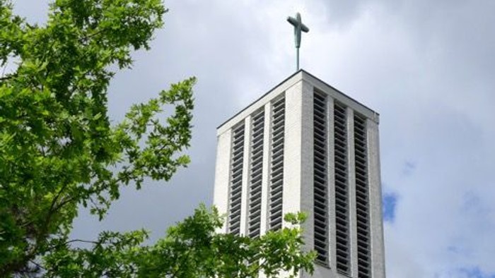 Moderner Kirchturm mit Kreuz, eingerahmt von üppigem grünem Laub unter einem bewölkten Himmel.