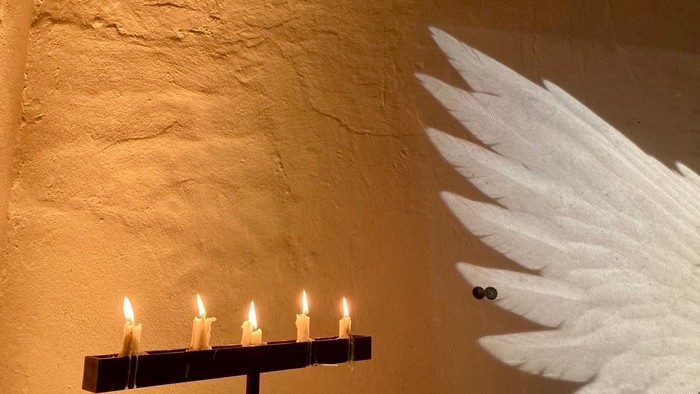Ein beleuchteter Kerzenhalter mit vier Kerzen neben einem dekorativen weißen Engel-Flügel aus Federn an einer strukturierten Wand.