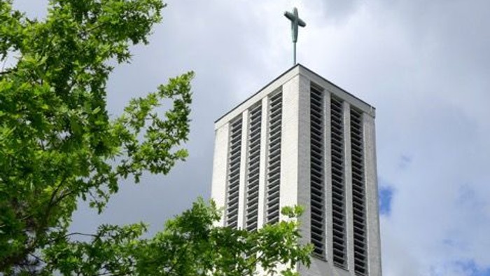 Moderner Kirchturm mit Kreuz, eingerahmt von üppigem grünem Laub unter einem bewölkten Himmel.