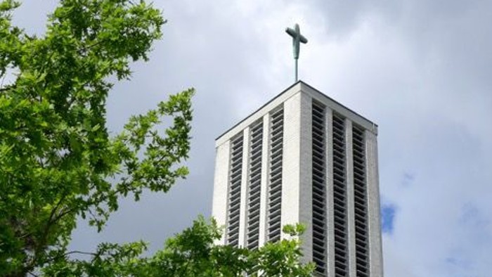 Moderner Kirchturm mit Kreuz, eingerahmt von üppigem grünem Laub unter einem bewölkten Himmel.