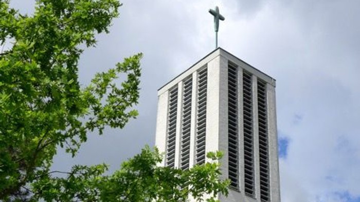 Moderner Kirchturm mit Kreuz, eingerahmt von üppigem grünem Laub unter einem bewölkten Himmel.