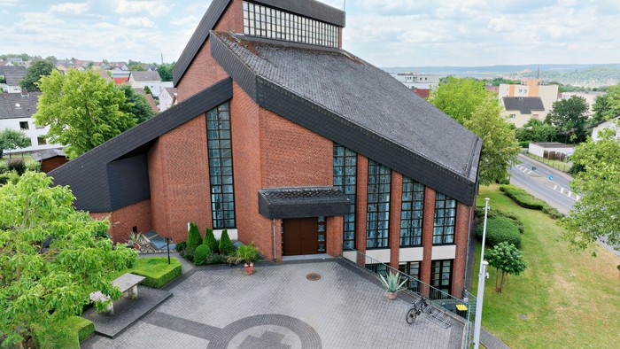 Das Bild zeigt eine moderne Kirche aus rotem Backstein mit großen Fenstern und einem gepflegten Vorhof.