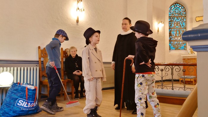 Børn i historiske kostumer leger i en dekoreret kirke sammen med en voksen.