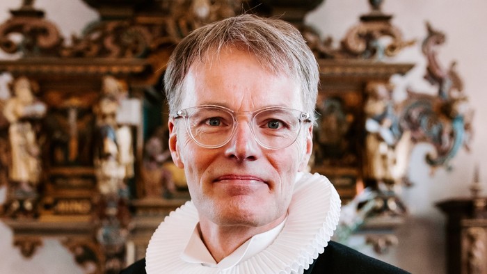 En mand i formelt gejstligt tøj med en hvid flip står i et rigt udsmykket rum.
