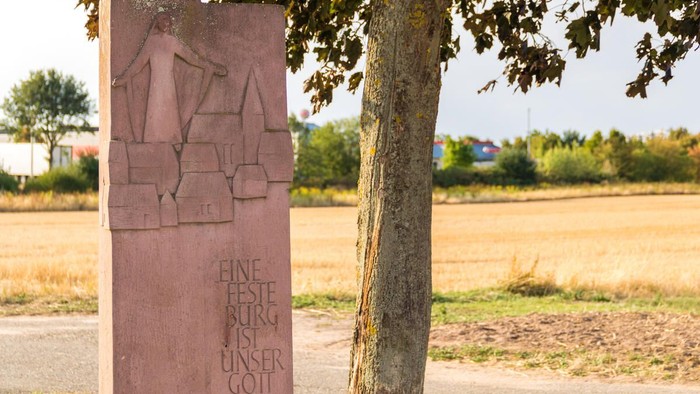 Sandsteinstele am Wegesrand mit reliefartiger Darstellung einer segnenden Christusfigur über stilisierten Häusern; darunter die Inschrift „Eine feste Burg ist unser Gott“. Daneben steht ein Baum, im Hintergrund ein abgeerntetes Feld und eine ländliche Lan