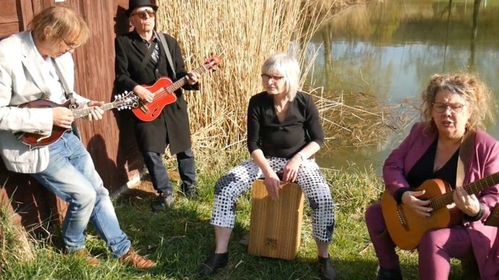 Vier Musiker*innen spielen gemeinsam am Ufer eines Sees neben einem kleinen Holzschuppen: eine Person mit Mandoline, eine mit E-Bass, eine auf einer Cajón sitzend und eine mit Akustikgitarre singend; im Hintergrund Schilf und Wasser, sonnige Atmosphäre im