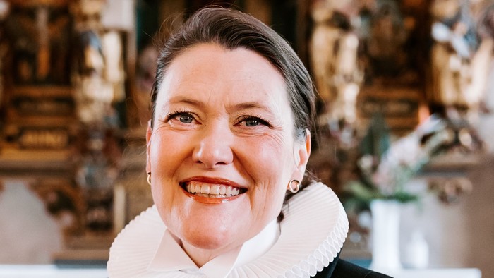 En kvinde i sort jakke med hvid krage står foran en prægtig altertavle.