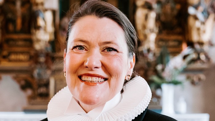 En kvinde i sort jakke med hvid krage står foran en prægtig altertavle.