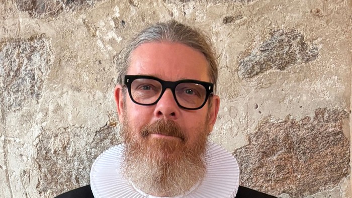 En mand i historisk tøj med en hvid kravekrøs, sort jakkesæt og briller poser foran en stenmur.