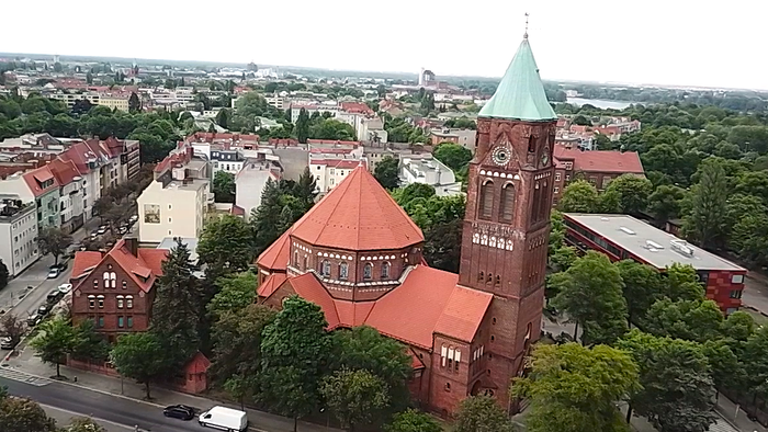 Luftaufnahme einer historischen Backsteinkirche mit hohem grünem Turm in einer grünen, städtischen Umgebung.