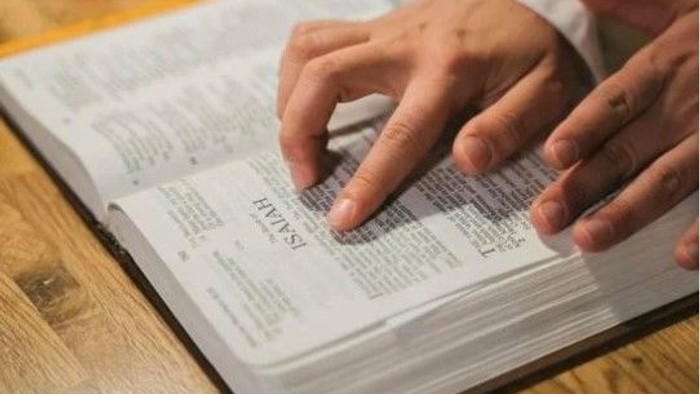 Eine Person, die in einem offenen Buch, wahrscheinlich einer Bibel, liest und auf den Text zeigt, auf einer hölzernen Oberfläche.