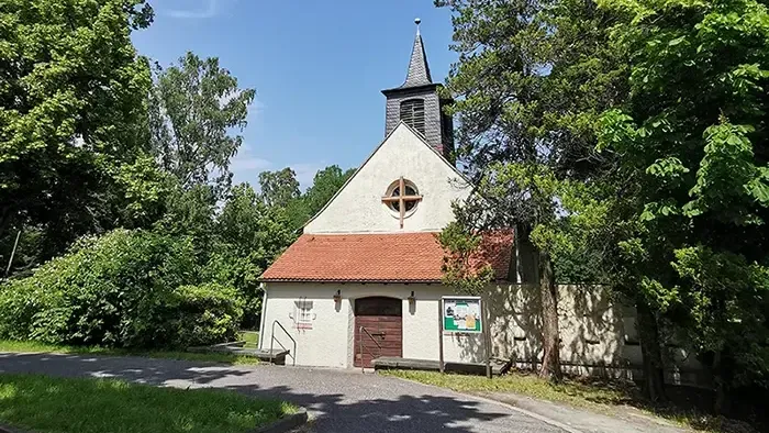 Kleine Kirche mit rotem Dach und Uhrturm, umgeben von Bäumen und Büschen.
