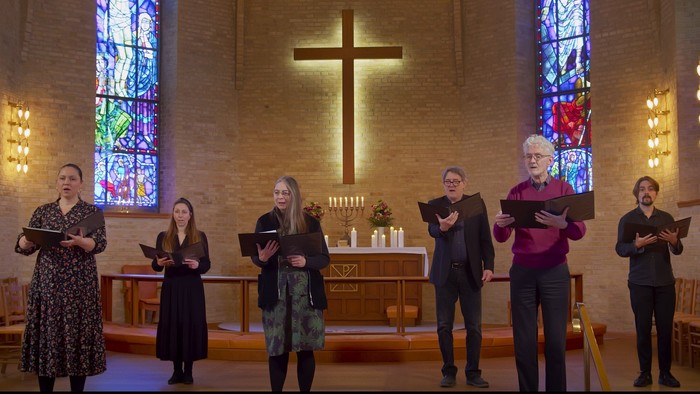 En gruppe mennesker, der synger eller læser fra salmebøger i en kirke med farvede glasvinduer og et stort kors.