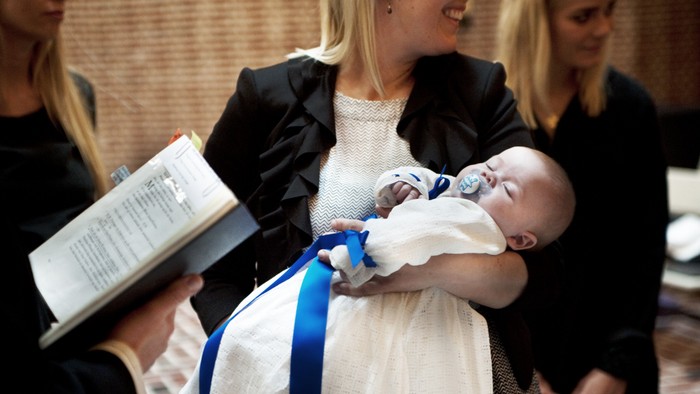 En kvinde holder et spæd barn, der er omvendt i en hvid kjole med blå bælte, mens en præst holder en bog.